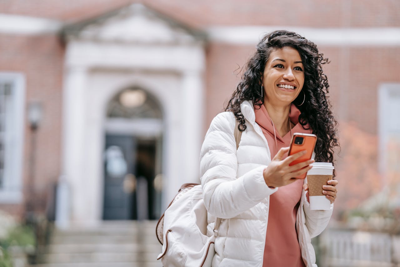 about-04 Cheerful young ethnic female student with long curly hair in casual clothes and backpack smiling and looking away while using mobile phone standing in campus with takeaway coffee